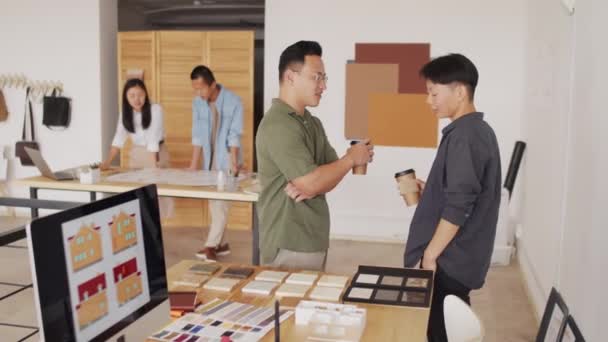 2人の若いアジア人建築家が職場でコーヒーブレイク中におしゃべりしながら 同僚たちが背景で建築計画を議論する様子を撮影した — ストック動画