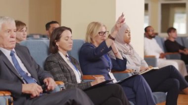 Orta yaşlı beyaz iş kadınının, büyük konferans salonundaki iş konferansında soru sormak için elini kaldırdığı orta boy bir fotoğraf.