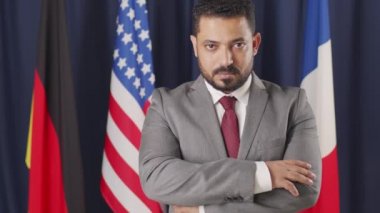 Orta Doğu ülkesinin ciddi orta yaşlı erkek siyasi liderinin orta boy portresi. Elleri katlanmış bir şekilde kameraya poz veriyor. Farklı ülkelerin bayrakları ile koyu mavi arka planda duruyor.