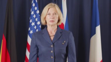 Orta yaşlı sarışın, beyaz kadın siyasi liderin koyu mavi arka planda kameraya poz veren orta yavaş portresi.