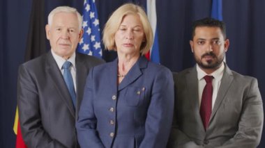 Birleşmiş Milletler bayraklarıyla koyu mavi arka planda duran kameraya bakan iki erkek ve bir kadın siyasi liderin orta yavaş portresini kaldır