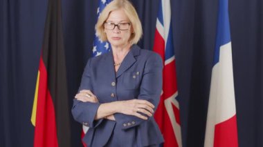 Kafkas kökenli orta yaşlı sarışın kadın politikacının koyu mavi arka planda BM bayraklarıyla kameraya poz veren orta yavaş portresi.