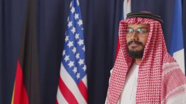 Geleneksel giysiler içinde genç Arap siyasi liderinin orta yavaş portresi ve koyu mavi arka planda kameraya poz vermesi.