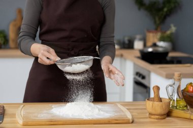 Önlüklü çağdaş kadın mutfak masasında un süzüyor.