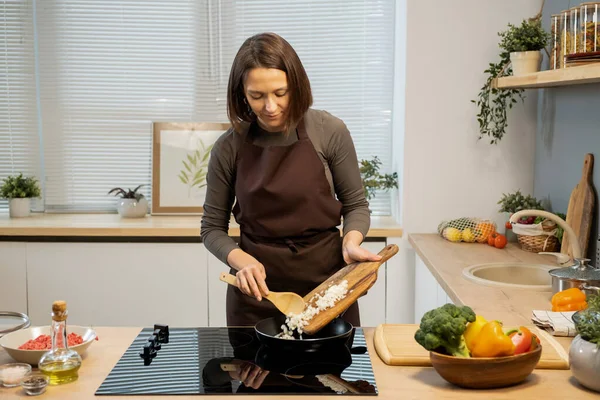 Young female in apron putting fresh chopped onion into frying pan — Foto Stock