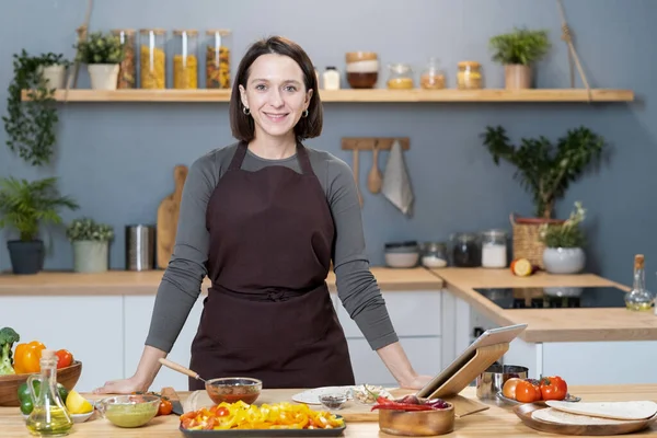 Young smiling female in apron standing by kitchen table — Foto Stock