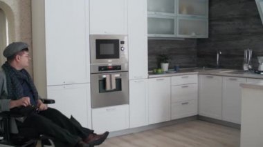 Long shot of Caucasian man in wheelchair entering his modern kitchen at home, turning on oven at daytime
