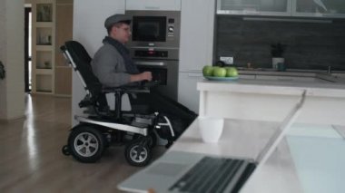 Low angle of Caucasian man in wheelchair wearing eyeglasses and peaked cap, entering kitchen, taking green apple from plate on cutting table