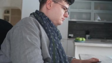 Side view of young man in electric wheelchair wearing eyeglasses sitting at desk at home at daytime, typing on portable computer