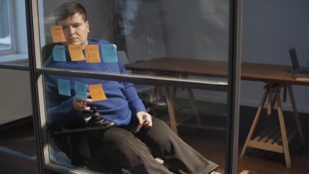 青い長袖の車椅子に身を包んだ白人男性のSteadicamは 夕方にオフィスのガラスの壁から粘着性のあるメモを取って 考えました — ストック動画