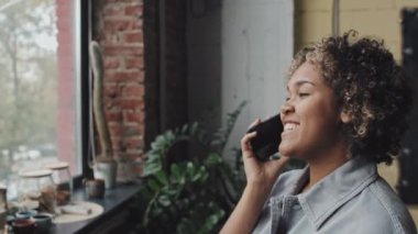 Genç siyahi bir kadının modern ofiste çerçeveye girmesi, cep telefonuyla konuşması, gülümsemesi.