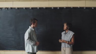 Beyaz adam ve genç kıvırcık siyah kadın gündüz vakti kara tahtanın önünde dikilip sohbet ediyorlar.