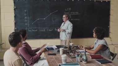 Yüksek açılı, gözlüklü, beyaz bir kadın profesör. Gündüz vakti masada oturan çeşitli öğrencilere karatahta grafiğini açıklıyor.