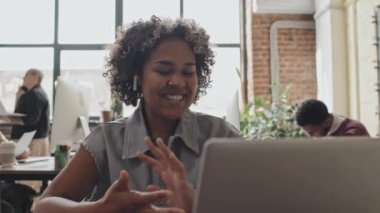 Kablosuz kulaklık takan neşeli, genç siyahi bir kadının göğsü. Gündüz vakti modern çalışma ortamında masada oturuyor. Taşınabilir bilgisayarla video görüşmesi.