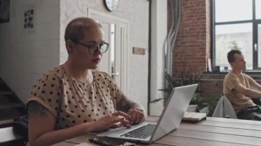 Gözlüklü, kısa saçlı, beyaz bir kadının gündüz vakti yoğun çalışma yerinde dizüstü bilgisayar kullanarak masada oturduğu orta ölçekli bir fotoğraf.