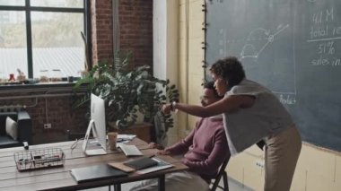 Orta uzunluktaki zeki, çift ırklı adam modern ofiste masada oturuyor, bilgisayar kullanıyor, genç siyahi kadın kenarda duruyor, konuşuyor ve tavsiyeler veriyor.