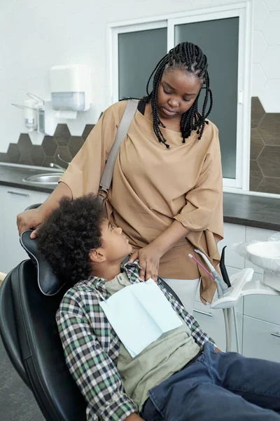 Ung afrikansk kvinna trösta sin lilla son sitter i fåtölj i tandläkare kontor — Stockfoto