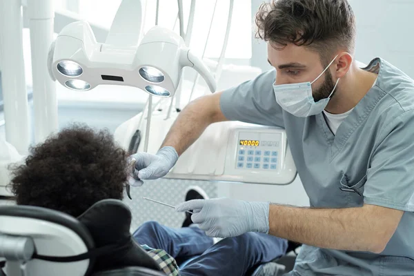 Dentista de uniforme, máscara e luvas segurando broca dentária e instrumento por boca de pequeno paciente — Fotografia de Stock