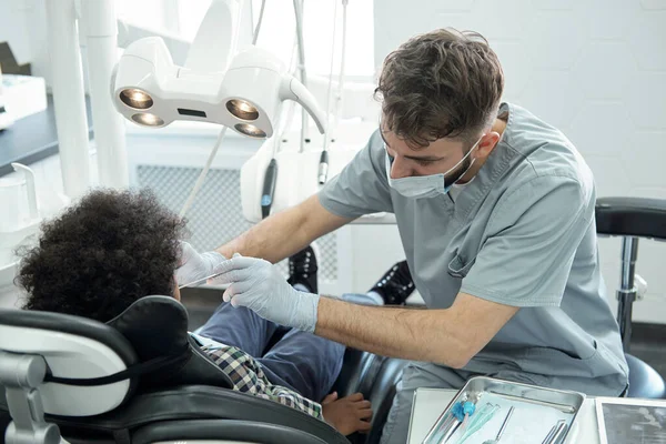 Dentist bending over little boy while drilling his tooth — Stock Photo, Image