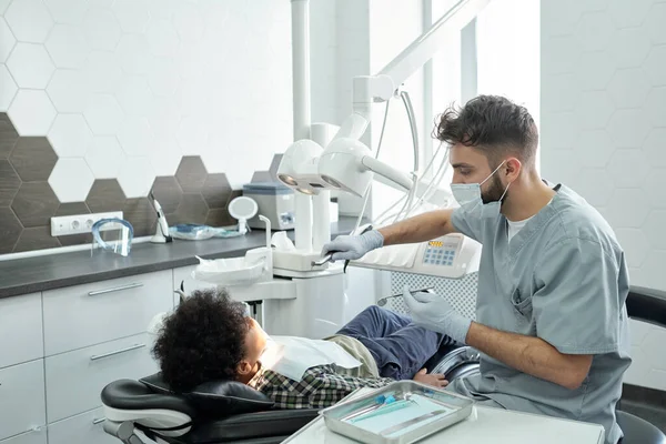 Сучасний стоматолог в робочому одязі збирається свердлити зуби маленького хлопчика — стокове фото