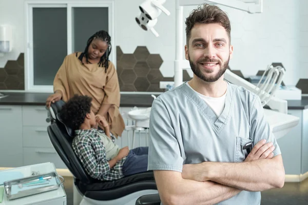 Jonge vrolijke tandarts kruising armen door borst tegen klanten — Stockfoto
