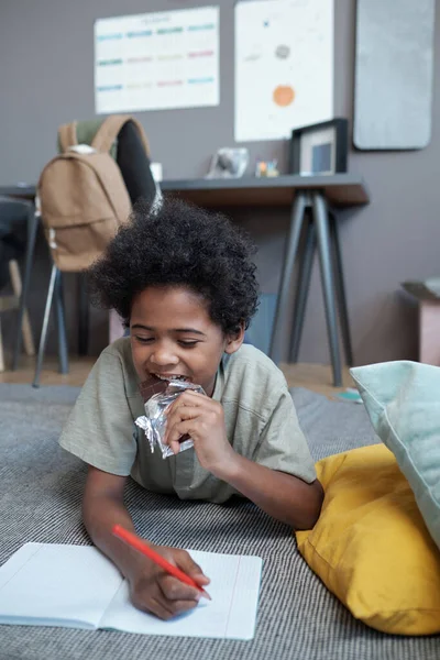 Niedlicher afrikanischer Junge isst Vollmilchschokolade, während er im Werbebuch zeichnet — Stockfoto