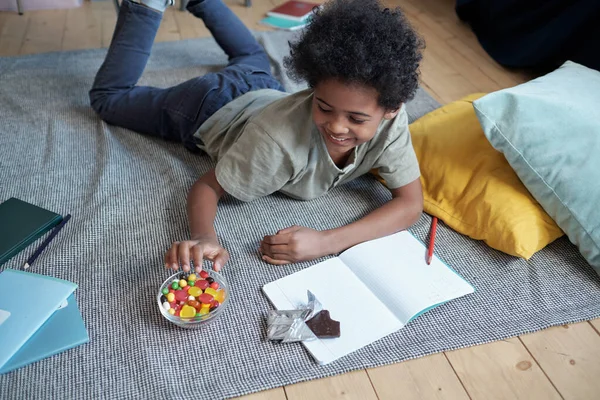 Mosolygó aranyos fiú vesz cukorkát ki az üvegtálból, miközben pihen a padlón — Stock Fotó
