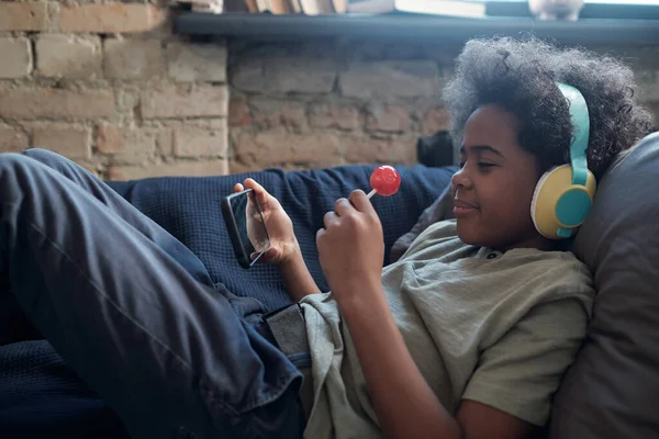 Happy cute African boy with lollipop and smartphone watching online movie — Stock Photo, Image