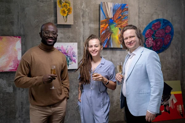 Drie gelukkige interculturele gasten van kunstgalerie met champagne — Stockfoto