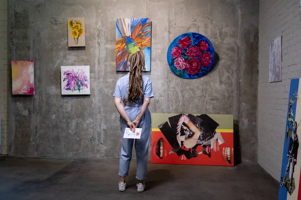 Visão traseira da jovem mulher em pé na frente da parede com coleção de obras de arte — Fotografia de Stock