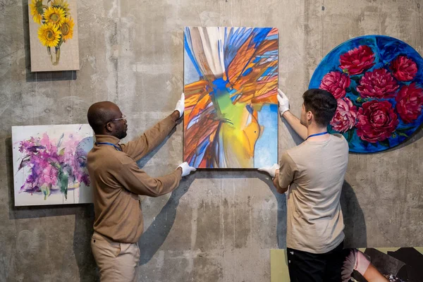Dois trabalhadores interculturais do sexo masculino de galeria pendurado pintura na parede — Fotografia de Stock
