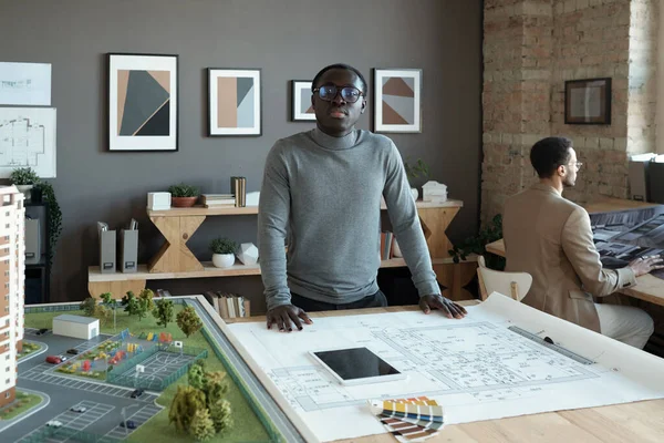 スケッチや家のレイアウトとテーブルのそばに立つ若いアフリカのエンジニア — ストック写真