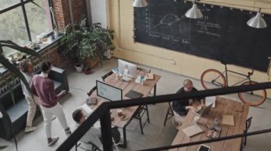 Gündüz vakti modern çalışma sahalarında yürürken konuşan melez erkek ve siyahi kadının yüksek açılı görüntüsü. Çalışma arkadaşları masalarda oturuyor.