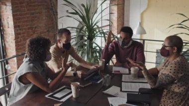 Yüz maskesi takan dört kadın ve erkek şirket çalışanının yüksek açılı görüntüsü. Gündüz vakti modern ofiste konferans masasında oturup iş görüşmesi yapıyorlar.