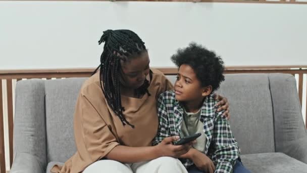 中等个子的黑人妇女和她十岁的儿子坐在接待室的沙发上 母亲用智能手机把儿子从发烧中引开 — 图库视频影像