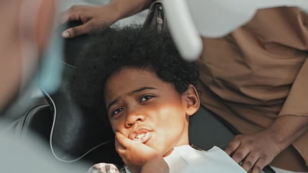 可爱的小学黑人男孩坐在牙医椅子上 与无法辨认的医生和养育儿子的父母交谈的特写镜头 — 图库视频影像