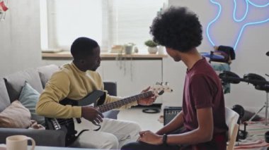 Oturma odasında kanepede oturan ve gündüz gitar çalan siyahi müzik öğretmenini izleyen genç adamın yan görüntüsü.