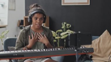 Gündüz vakti modern dairede koltukta oturan, sentezleyici çalan, el sallayan, gülümseyen, konuşan ve kamerayla çekim yapan 11 yaşındaki siyahi kız çocuğu.