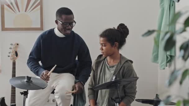 中程度の長さの準備黒男性チューター身に着けている眼鏡 教える11歳アフリカ系アメリカ人の女の子使用ドラム上の電子ドラムで現代的なアパートで昼間 — ストック動画