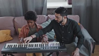 Orta boylu, koyu saçlı, Asyalı bir erkek öğretmen. Küçük zenci çocuğa evde kanepede otururken sentezleyici çalmasını öğretiyor.