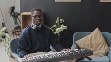 Boynuna gözlük takan, kulaklık takan, modern bir dairede oturan, sentezleyiciyle poz veren, kameraya bakıp gülümseyen çömez siyah bir adamın orta boy portresi.