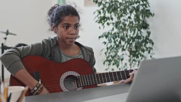 11歳の黒の少女の中間クローズアップは 昼間の現代のアパートで彼女の部屋のポータブルコンピュータを介してオンラインアコースティックギターのレッスンを持っています — ストック動画