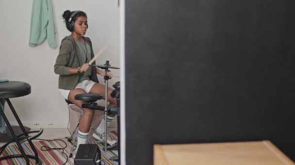 才能のある11歳のアフリカ系アメリカ人の少女のフルショットは 日中に彼女の部屋のモダンなアパートで電子ドラムを楽しんで 耳の上のヘッドフォンを身に着けて — ストック動画