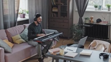 Asyalı bir adamın sakal ve kulaklık takıp sentezleyicinin önündeki koltukta otururken, konuşurken ve gülümserken gündüz vakti taşınabilir bilgisayardan video araması.