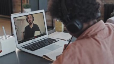 Kulaklıklı, siyah kıvırcık saçlı, kulaklık takan, öğleden sonra masasında oturan, taşınabilir bilgisayarda Asyalı erkek öğretmenle online ders alan siyah kıvırcık çocuk.