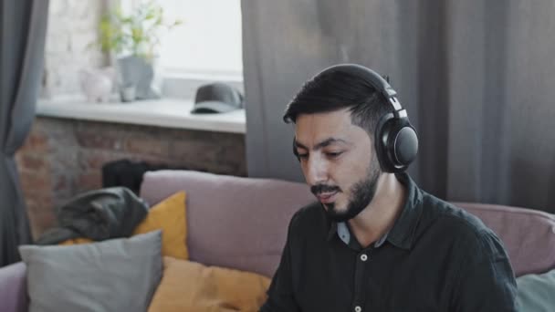 Lehajolt Fiatal Ázsiai Férfi Szakállal Fülhallgatóval Kanapén Otthon Szintetizátorozik Beszél — Stock videók