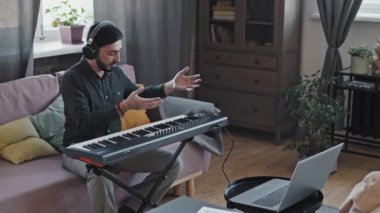 Orta uzunlukta sakal ve kulaklık takan Asyalı bir adam kanepede oturuyor, sentezleyici oynuyor, konuşuyor ve gülümsüyor, portatif bilgisayardan gündüz vakti video çağrısı yapıyor.