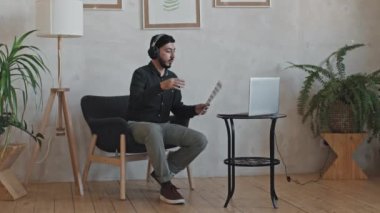 Asyalı erkek müzik öğretmeninin sakallı ve kulaklıklı bir şekilde evdeki koltukta otururken çevrimiçi ders verirken bilgisayar kamerasında notlar gösteren bir fotoğrafı.