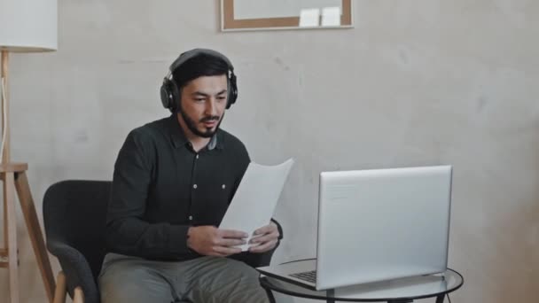 午後には自宅の椅子に座って髭や耳以上のヘッドフォンを身に着けているアジアの教師の中長期 ポータブルコンピュータを使用してオンラインレッスンを教えて — ストック動画