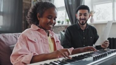 Asyalı profesyonel erkek müzik öğretmeninin elinde notalarla düşük açılı notalar, sentezleyicide beste yapan, gülümseyen, evdeki kanepede oturan on yaşında siyah bir kız.
